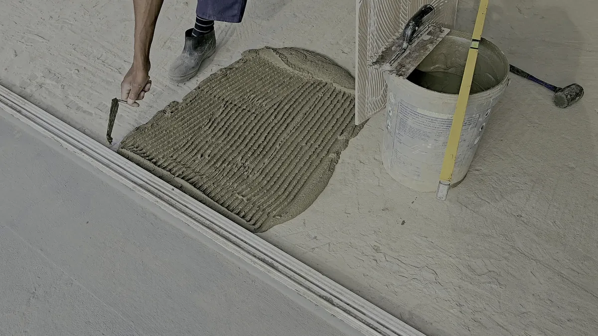 Handwerker rührt Fliesenkleber im Eimer an, daneben Zahnkelle, Wasserwaage und angelehnte Fliese vor gekämmtem Kleberbett